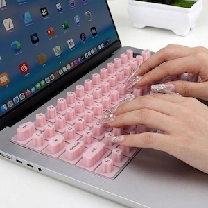 Può includere: Un computer portatile con una copertura per tastiera in silicone rosa chiaro, caratterizzata da tasti rialzati di forma cilindrica. La copertura si sovrappone ai tasti standard del portatile e presenta lettere e numeri stampati in nero.