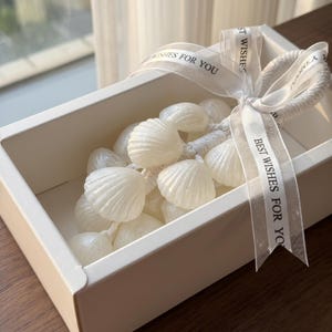 May include: A white gift box filled with white seashell-shaped ornaments. A white ribbon with the text "BEST WISHES FOR YOU" is tied around the box. The ornaments are strung together with a white cord.