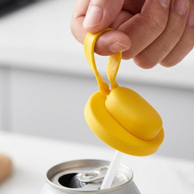 May include: A bright yellow drink can lid with a loop handle, designed to fit over a can and accommodate a straw. The lid is shown being held by a hand, with a silver can and white straw visible.