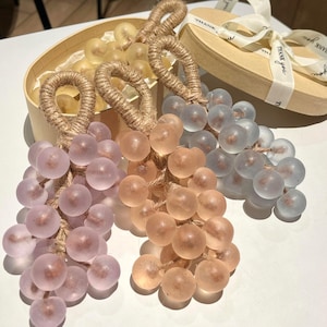 Puede incluir: Racimos de uvas decorativos en tonos lavanda, melocotón y transparente. Cada racimo presenta cuentas redondas translúcidas y un lazo de cuerda de yute. Algunos se presentan en una caja de regalo redonda, atados con una cinta.