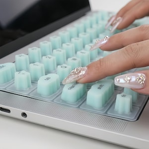 May include: A light mint green silicone keyboard cover featuring uniquely shaped, scalloped keys with black printed text including tab, caps lock, and shift. The cover is placed over a silver laptop keyboard, providing a textured, tactile surface.