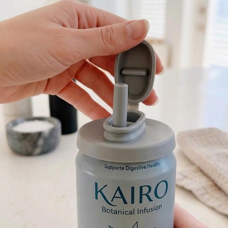 May include: A light gray bottle with a matching lid and straw. The bottle has the word "KAIRO" in large letters, with "Botanical Infusion" below. The text "Supports Digestive Health" is also visible. The bottle is being opened.