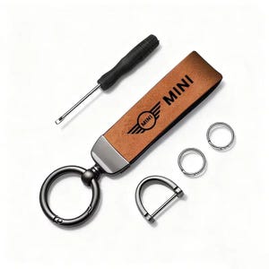 May include: A brown leather key fob with the word "MINI" and logo, a screwdriver, two key rings, and a D-ring. The key fob has a metal accent and a large key ring. The items are arranged on a white background.