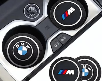 May include: Black car cup coasters with white trim and BMW and M logos in blue, white, and red. The coasters are shown in a car's center console, with two additional coasters visible.