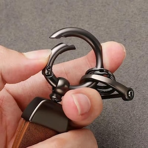 May include: A close-up of a gunmetal-colored keychain clasp. The clasp is open, revealing the hook and the spring-loaded mechanism. The keychain has a brown leather strap attached to it.