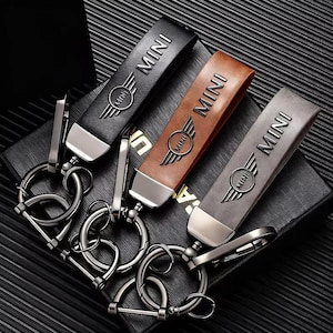 May include: Three leather keychains in black, brown, and gray, each embossed with the word "MINI" and a winged emblem. The keychains have silver-toned metal hardware, including key rings and clasps. The keychains are displayed on a dark surface.