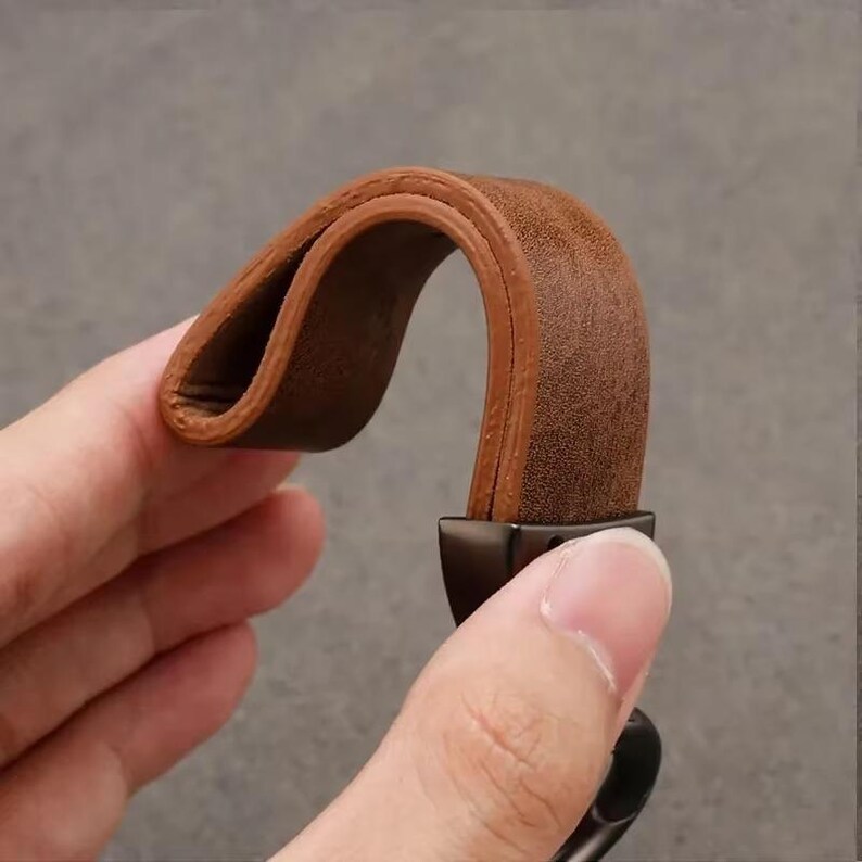 May include: A close-up of a brown leather strap, bent into an arch. The strap is a rich, warm brown color with a textured surface. The strap is attached to a black metal clasp. The image is set against a neutral gray background.