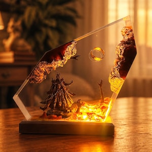 Könnte beinhalten: Eine dekorative Resin-Skulptur mit einer Szene mit einem goldenen Ring, einer dunklen Figur und einer feurigen Landschaft. Die Skulptur ist in einem klaren, abgewinkelten Block mit einem Holzsockel eingeschlossen. Die Szene wird von warmem, gelbem Licht beleuchtet.