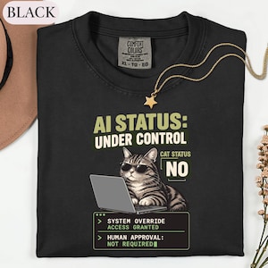 May include: A black t-shirt featuring a graphic of a tabby cat wearing sunglasses while using a laptop. The text reads AI STATUS: UNDER CONTROL, CAT STATUS NO, SYSTEM OVERRIDE ACCESS GRANTED, and HUMAN APPROVAL: NOT REQUIRED. The shirt has a Comfort Colors label.