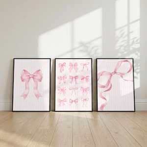 May include: Three framed wall art prints featuring watercolour illustrations of pink bows on a white background with thin vertical pink stripes. The left print shows one large bow, the centre shows twelve small bows, and the right shows one large ribbon.