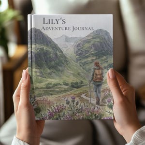 May include: A hardback journal titled Lily's Adventure Journal featuring a watercolour illustration of a person with a backpack and hiking pole walking on a path through a green mountain valley with purple wildflowers in the foreground.