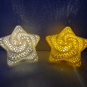 May include: Two star-shaped lamps with an intricate, open-work geometric lattice pattern. One lamp emits a cool white light, while the other emits a warm yellow light. Both objects feature a hollow, three-dimensional structure with cutouts.