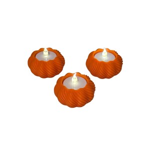 May include: Three orange tea light holders with a textured, spiraled exterior design. Each holder contains a small, white, battery-operated LED candle with a glowing flame-shaped tip, providing a soft light from the center of the orange base.