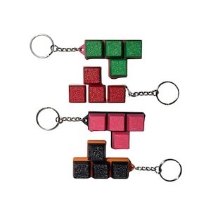 Puede incluir: Cuatro llaveros con formas de bloques geométricos inspirados en juegos de rompecabezas clásicos. El conjunto incluye una forma de T verde, una forma de L roja, una forma de T rosa y una forma de bloque naranja y negro, cada una unida a una anilla de metal plateado.