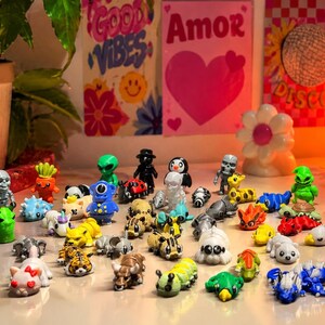 Puede incluir: Una colección de coloridas figuras en miniatura, que incluyen extraterrestres, animales y monstruos, dispuestas sobre una superficie. Las figuras son de varios colores, como verde, azul y amarillo. El fondo incluye carteles con las palabras "Good Vibes" y "Amor".