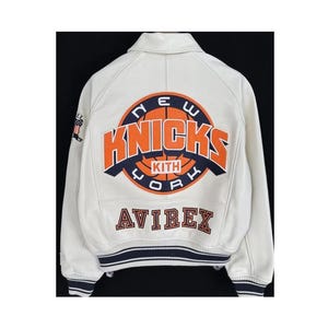 May include: A white leather varsity jacket featuring a large circular New York Knicks logo on the back with orange and navy blue lettering. The text KITH is centered within the basketball graphic, and the word AVIREX is embroidered below in navy.