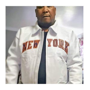 May include: A white zip-up jacket featuring a classic collar and long sleeves. The text NEW YORK is embroidered across the chest in bold, arched lettering with dark blue and orange outlines, layered over a solid black shirt underneath.