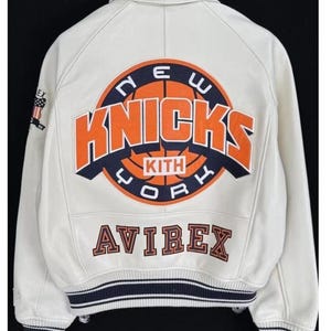 May include: A white leather varsity jacket featuring a large circular graphic on the back with the text NEW YORK KNICKS and KITH in orange, navy blue, and white. The word AVIREX is embroidered below in navy blue and orange. Navy blue striped ribbed trim.