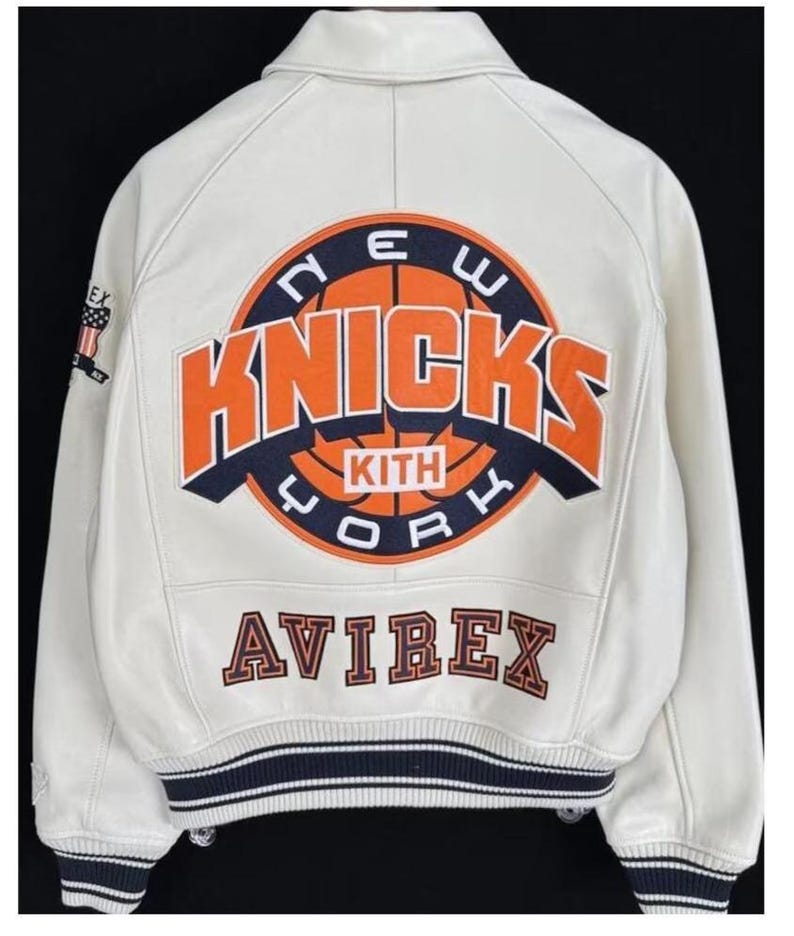 May include: A white leather varsity jacket featuring a large circular graphic on the back with the text NEW YORK KNICKS and KITH in orange, navy blue, and white. The word AVIREX is embroidered below in navy blue and orange. Navy blue striped ribbed trim.
