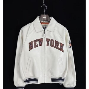 May include: A white leather bomber jacket featuring a full-length front zipper, a classic collar, and ribbed cuffs and hem with navy blue stripes. The chest displays the text NEW YORK in large, dark-outlined letters, with an American flag patch on the sleeve.