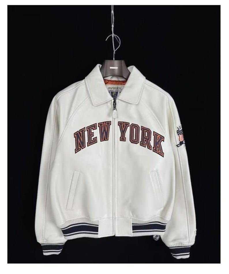 May include: A white leather bomber jacket featuring a full-length front zipper, a classic collar, and ribbed cuffs and hem with navy blue stripes. The chest displays the text NEW YORK in large, dark-outlined letters, with an American flag patch on the sleeve.