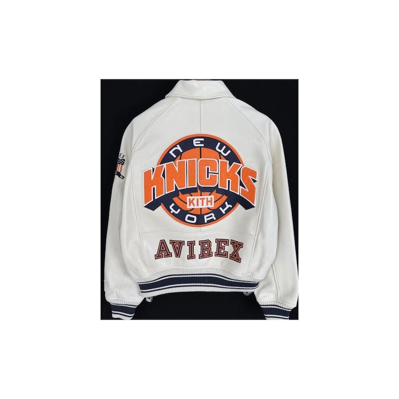 May include: A white leather varsity jacket featuring a large circular New York Knicks logo on the back with orange and navy blue lettering. The text KITH is centered within the basketball graphic, and the word AVIREX is embroidered below in navy.