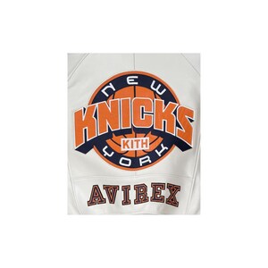 May include: A white leather jacket featuring a large circular graphic on the back. The graphic displays a basketball with the text NEW YORK KNICKS in orange and navy blue, with KITH in the center. The word AVIREX is embroidered below in navy.