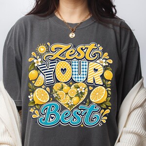 May include: A charcoal gray t-shirt featuring a graphic print with the text Zest Your Best in yellow, blue, and white. The design includes illustrations of yellow lemons, green leaves, white blossoms, and a heart shape filled with lemon slices.