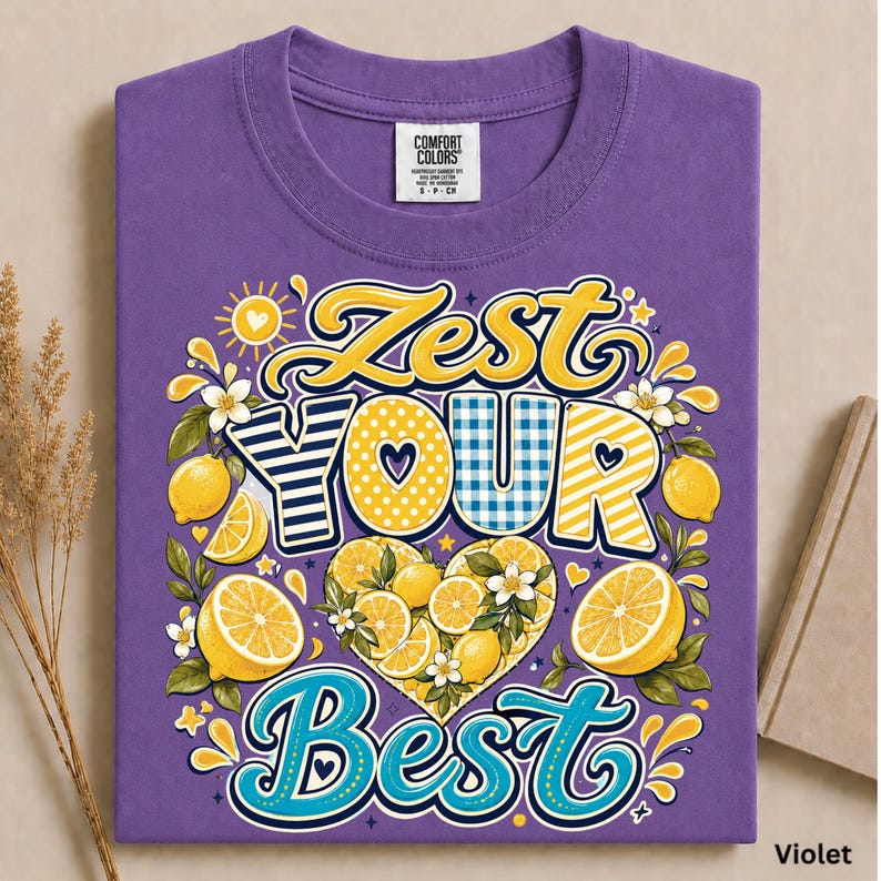 May include: A violet t-shirt featuring a graphic with the text ZEST YOUR BEST. The design includes yellow lemons, white blossoms, and a heart-shaped arrangement of lemon slices. The lettering uses yellow, blue, and white patterns like stripes.
