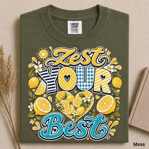 May include: An olive green t-shirt featuring a graphic print with the text ZEST YOUR BEST. The design includes yellow lemons, white blossoms, and decorative patterns in yellow, blue, and white stripes and polka dots arranged in a heart shape.