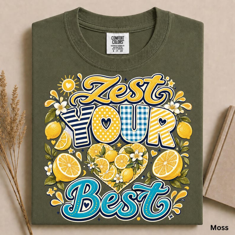May include: An olive green t-shirt featuring a graphic print with the text ZEST YOUR BEST. The design includes yellow lemons, white blossoms, and decorative patterns in yellow, blue, and white stripes and polka dots arranged in a heart shape.