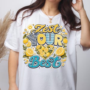 May include: A white t-shirt featuring a graphic design with the text Zest Your Best in blue and yellow lettering. The design includes illustrations of yellow lemons, green leaves, white flowers, and a heart shape filled with lemon slices.