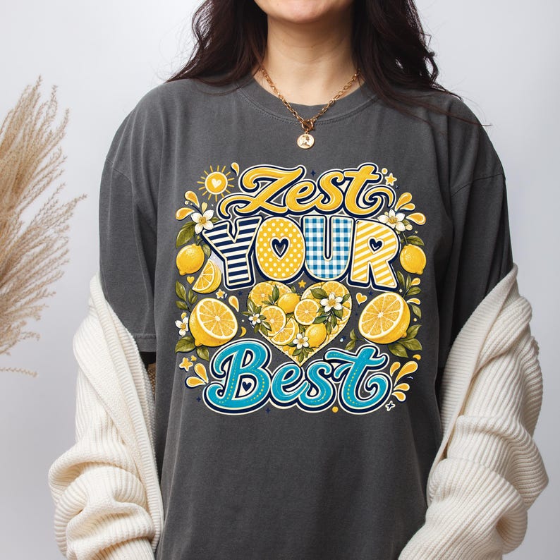 May include: A charcoal gray t-shirt featuring a graphic print with the text Zest Your Best in yellow, blue, and white. The design includes illustrations of yellow lemons, green leaves, white blossoms, and a heart shape filled with lemon slices.