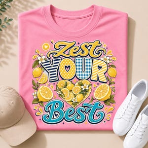 May include: A pink t-shirt featuring a graphic design with the text ZEST YOUR BEST in yellow, blue, and white. The design includes illustrations of yellow lemons, green leaves, and white flowers arranged in a heart shape with patterns.