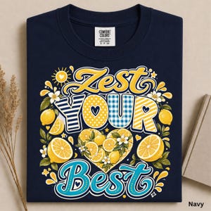 May include: A navy blue t-shirt featuring a graphic design with the text "Zest Your Best" in yellow and blue lettering. The design includes illustrations of yellow lemons, green leaves, white blossoms, and a central heart shape filled with lemon slices.