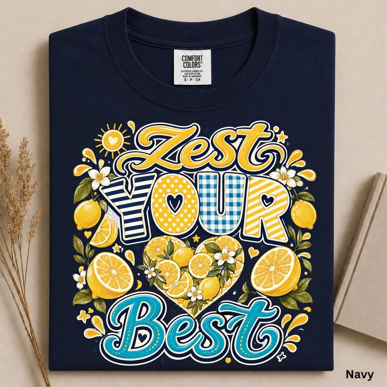 May include: A navy blue t-shirt featuring a graphic design with the text "Zest Your Best" in yellow and blue lettering. The design includes illustrations of yellow lemons, green leaves, white blossoms, and a central heart shape filled with lemon slices.