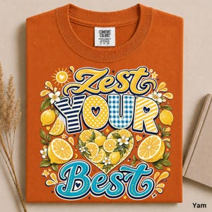 May include: A burnt orange t-shirt featuring a graphic print with the text Zest Your Best. The design includes yellow lemons, green leaves, white blossoms, and a heart shape filled with citrus slices, all set against a sunburst background.