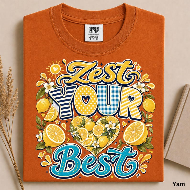 May include: A burnt orange t-shirt featuring a graphic print with the text Zest Your Best. The design includes yellow lemons, green leaves, white blossoms, and a heart shape filled with citrus slices, all set against a sunburst background.