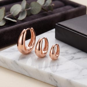 May include: Three rose gold hoop earrings of varying sizes arranged in a row. Each earring features a smooth, polished, thick, rounded design with a hinged closure mechanism. The hoops are displayed in descending order from largest to smallest.