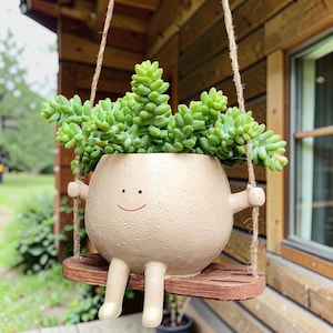 Puede incluir: Una maceta de cerámica beige, redonda y texturizada, con una cara sonriente sencilla, dos brazos y dos piernas. La maceta descansa sobre un pequeño columpio de madera rectangular marrón, suspendido por dos cuerdas de yute, con una suculenta.