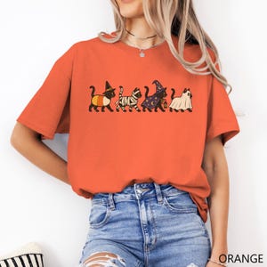 May include: An orange short-sleeved t-shirt featuring a graphic print of four black cats in Halloween costumes. The cats are dressed as a candy corn, a mummy, a wizard with a star-patterned hat and cloak, and a ghost wearing a white sheet.