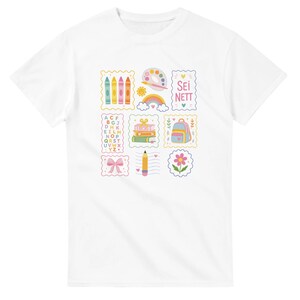 Puede incluir: Una camiseta blanca de manga corta con un estampado gráfico central de varios iconos escolares coloridos dispuestos en una cuadrícula, que incluyen ceras, una paleta de pintura, un arcoíris, libros, una mochila, un lápiz, una flor y el texto SEI NETT.