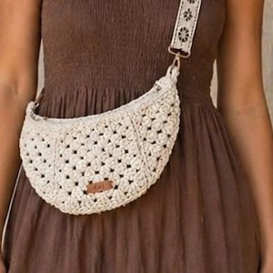 May include: A crescent-shaped off-white crochet crossbody bag with a textured granny square pattern and a small brown rectangular brand tag. The bag features a wide, cream-coloured woven shoulder strap decorated with a repeating floral pattern.