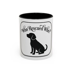 May include: White ceramic mug with a black interior and rim. The mug features a black silhouette of a dog and the text "Who Rescued Who?" in a decorative font, all within a rectangular frame.
