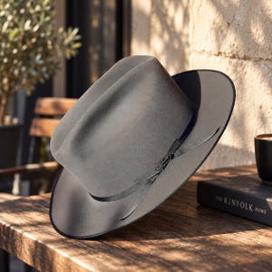 Op de afbeelding: Een grijze vilten fedora hoed met een brede rand en een ingedeukte kroon. De hoed heeft een dunne zwarte lintband rond de basis van de kroon, geaccentueerd met een klein metalen detail. De rand is afgewerkt met een zwarte bies.