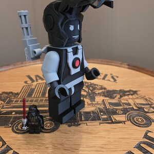 Peut inclure: Une figurine de style Lego imprim&eacute;e en 3D, avec un sch&eacute;ma de couleurs noir et gris, pr&eacute;sentant un design de casque unique et un bras m&eacute;canique. Une figurine Lego Darth Vader plus petite avec un sabre laser rouge est &eacute;galement pr&eacute;sente. Les figurines sont expos&eacute;es sur une surface en bois.