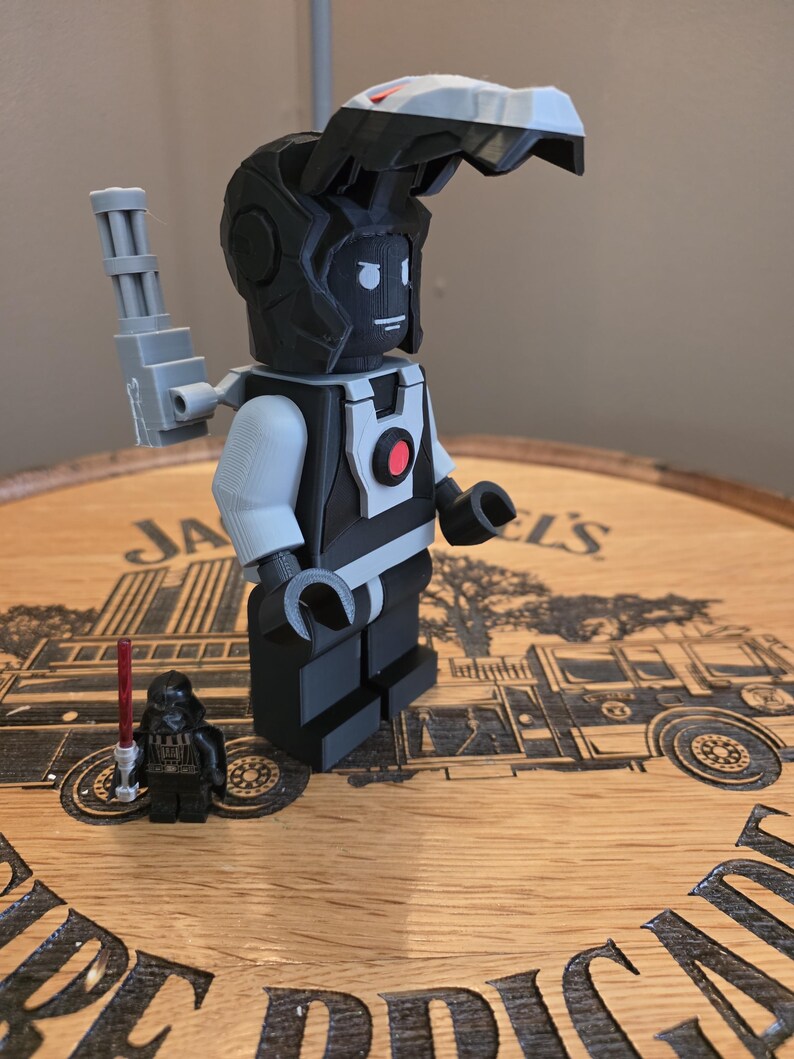 Peut inclure: Une figurine de style Lego imprim&eacute;e en 3D, avec un sch&eacute;ma de couleurs noir et gris, pr&eacute;sentant un design de casque unique et un bras m&eacute;canique. Une figurine Lego Darth Vader plus petite avec un sabre laser rouge est &eacute;galement pr&eacute;sente. Les figurines sont expos&eacute;es sur une surface en bois.