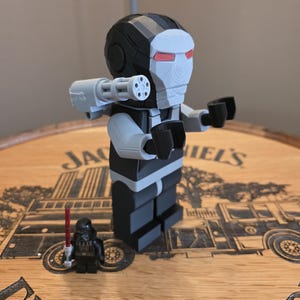 Peut inclure: Une figurine de War Machine noire, grise et blanche avec un canon mont&eacute; sur l'&eacute;paule et des d&eacute;tails d'yeux rouges. Une petite figurine de Dark Vador avec un sabre laser rouge est &eacute;galement pr&eacute;sente. Les figurines sont sur une surface en bois.