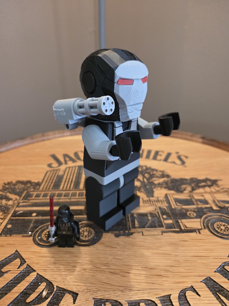 Peut inclure: Une figurine de War Machine noire, grise et blanche avec un canon mont&eacute; sur l'&eacute;paule et des d&eacute;tails d'yeux rouges. Une petite figurine de Dark Vador avec un sabre laser rouge est &eacute;galement pr&eacute;sente. Les figurines sont sur une surface en bois.