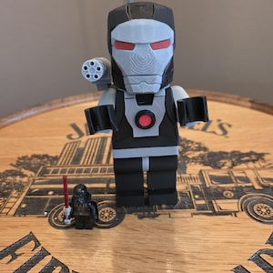 Peut inclure: Une figurine grise et noire de War Machine avec des d&eacute;tails rouges sur les yeux et un canon mont&eacute; sur l'&eacute;paule. Une petite figurine de Dark Vador avec un sabre laser rouge est &eacute;galement pr&eacute;sente. Les figurines sont sur une surface en bois.